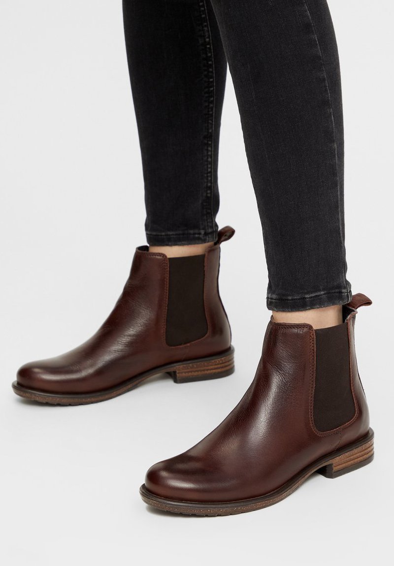 Botas de cuero marrón hasta el tobillo con paneles laterales elásticos, punteras redondas y tacones de madera apilados, combinadas con jeans ajustados negros.