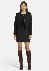 Veste courte gris foncé avec deux poches avant, associée à une mini-jupe noire ajustée et des bottes marron brillant jusqu'aux genoux avec un motif texturé.