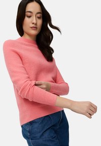 Rosa gerippter Strickpullover mit Rundhalsausschnitt und langen Ärmeln, kombiniert mit blauen Jeans, vor weißem Hintergrund dargestellt.