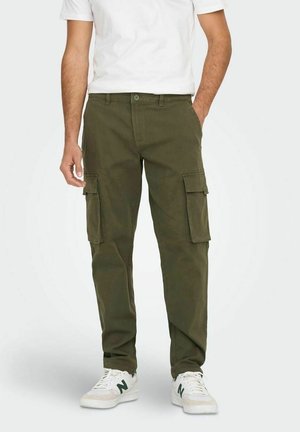 Mand iført olivengrønne cargobukser med klaplommer, en hvid t-shirt og hvide sneakers med mørkegrøn logo, stående mod en ensfarvet baggrund.