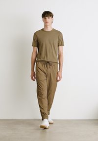 Olivgrünes kurzärmeliges Hemd und hellbraune elastische Bundhose aus strukturiertem Stoff, kombiniert mit weißen Sneakern vor einem neutralen Hintergrund.