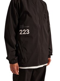 Sort lynlåstrøje med "223" og lille tekst trykt i hvid på venstre side, båret over en hvid skjorte og sorte bukser.