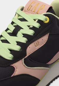 Μαύρο και ροζ αθλητικό παπούτσι με φόντο νέον πράσινα κορδόνια, εμφανίζοντας το λογότυπο "GAP" στη γλώσσα και στη πλευρά κοντά στις οπές των κορδονιών.