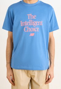 Světle modré bavlněné tričko s krátkými rukávy, na kterém je růžový nápis "The Intelligent Choice" a malá značka New Balance na boku.
