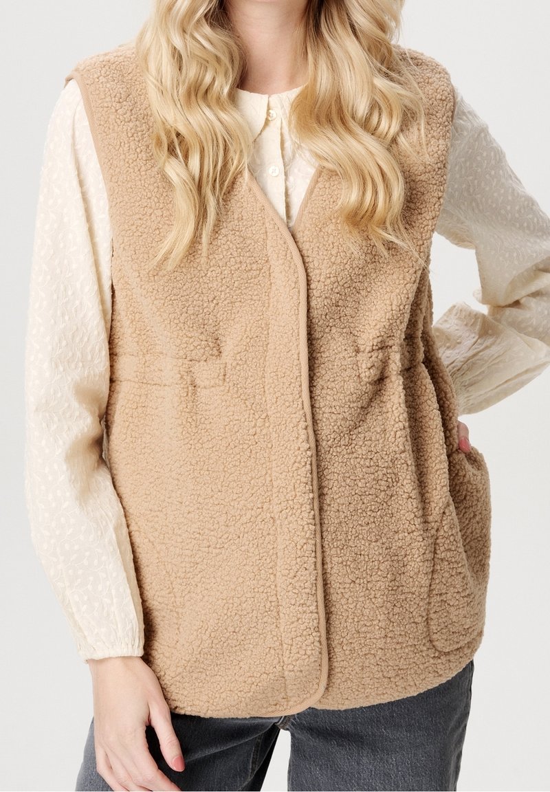 Veste en polaire sherpa beige portée sur un chemisier crème à manches longues avec un motif texturé et un pantalon gris foncé, main reposant dans la poche de la veste.