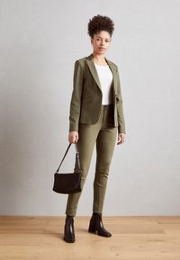 Olivově zelený střižený blazer a kalhoty, přiléhavý bílý top, černá kabelka a černé holínkové boty na podpatku. Hladká tkanina s propracovaným designem.