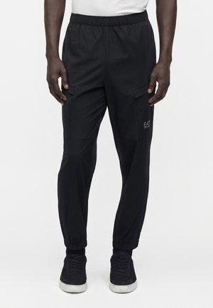 Bărbat purtând pantaloni cargo negri marca EA7 cu manșete elastice și adidași negri, stând pe un fundal deschis.