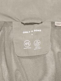 Ετικέτα ρούχου με την επωνυμία "ONLY & SONS", μέγεθος L, με εικονίδια αντιανεμικής και αδιάβροχης προστασίας σε γκρι διχτυωτό ύφασμα.