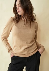 Beige neule pyöreällä kauluksella, ribatut hihat ja helma. Valmistettu pehmeästä, sileästä materiaalista. Yhdistetty mustiin housuihin.
