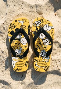 Tongs jaunes Havaianas avec des brides noires présentant des illustrations en noir et blanc de Mickey Mouse et ses amis sur un sol sablonneux.