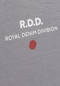 Pelēka kokvilnas T-krekls ar baltu tekstu "R.D.D. ROYAL DENIM DIVISION" un nelielu sarkanu apļa akcentu. Gluda tekstūra, ikdieniša dizains.