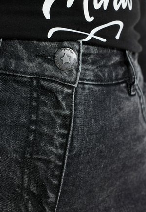 Czarne jeansy z denimu z lekkim połyskiem, z metalowym guzikiem z motywem gwiazdy i fakturowaną tkaniną. Widok z bliska na obszar pasa.