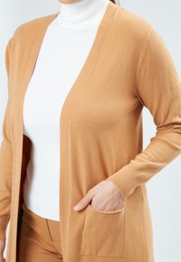 Een lange cardigan gemaakt van zacht materiaal, met een sjawlkraag, open voorkant, lange mouwen en twee voorzakken. Draagt over een witte coltrui.