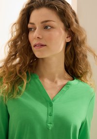 Grön bomullsskjorta med V-ringning och tre knappar. Tyget är slätt med en lätt lyster. Modellen har vågigt hår och vänder sig åt vänster.