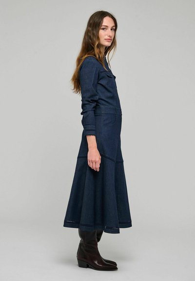 Femme aux longs cheveux bruns portant une longue robe en denim bleu foncé et des bottes noires mi-mollet, debout de profil sur un fond uni.