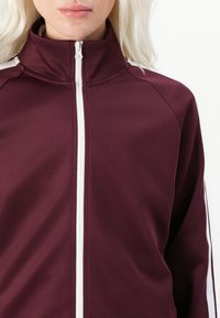 Burgundin värinen vetoketjullinen takki, jossa on korkea kaulus ja valkoiset taitteet sivuilla. Suunnistus on sileä ja urheilullinen. Vetoketju kulkee keskellä.