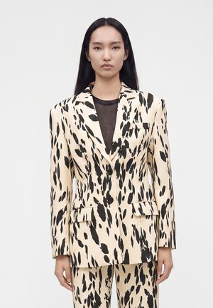 Femme aux longs cheveux noirs et raides, portant un costume crème avec des taches noires irrégulières et un top noir transparent en dessous, debout devant un fond uni.
