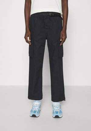 Personne portant un pantalon cargo noir ample avec des poches latérales, un haut blanc et des baskets bleu clair et blanc, debout devant un fond uni.