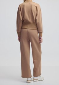 Persona che indossa un felpa beige e pantaloni a gamba larga abbinati, in piedi con la schiena rivolta alla fotocamera su uno sfondo chiaro e semplice.