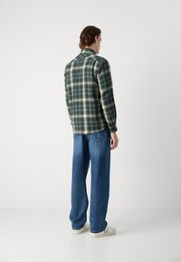 Checked green and yellow flannel shirt with a pointed collar and long sleeves, paired with loose-fit blue jeans and white sneakers.