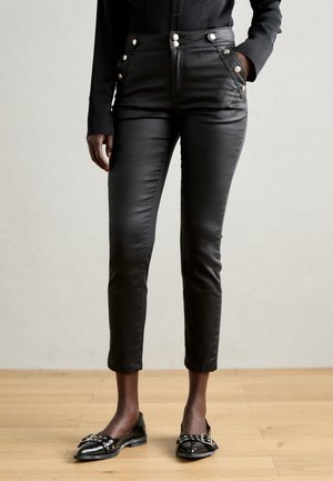Zwarte slim-fit broek met gouden knopen op zakken en taille, gedragen met een zwart overhemd en zwarte platte schoenen met gesp en studs.