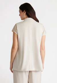 Jasno beżowy bezrękawnik z wyraźnym kołnierzem, gładką tkaniną i wzorzystym akcentem na ramieniu. Posiada prosty tył i subtelny połysk.