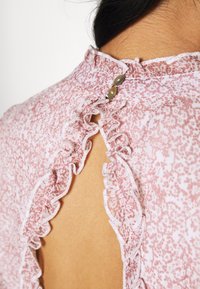 Rückansicht einer Person, die eine rosa Blumenshirt mit Rüschenbesatz trägt und zwei kleine Knöpfe hat, die eine Schlüssellochöffnung am Hals verschließen.