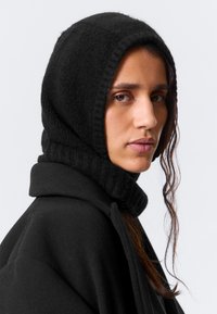 Capuz preto de malha com bordas caneladas, combinado com um casaco preto. Apresenta um acabamento texturizado e um design ajustado à volta do rosto.