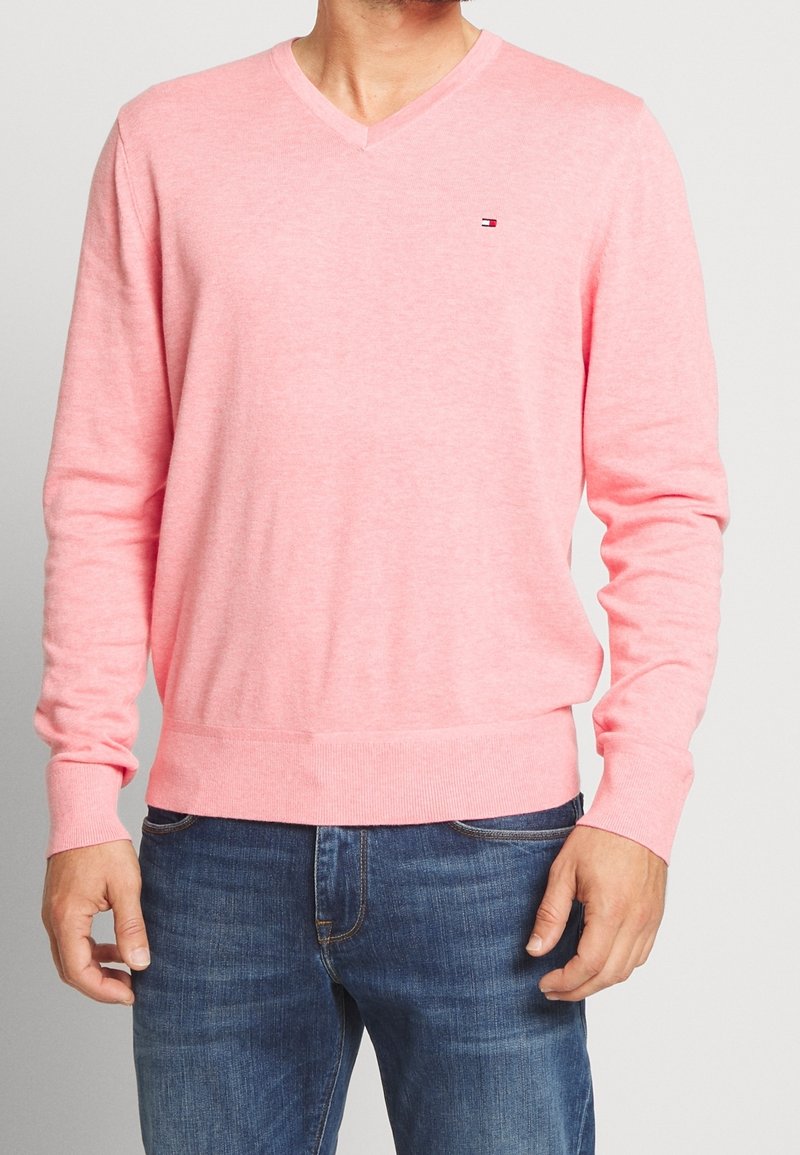 Homme portant un pull rose clair à manches longues avec col en V et un jean bleu, debout devant un fond clair uni.