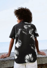 Svart kortärmad skjorta med vita palmbladsmönster. Lätt tyg med krage och avslappnad passform.
