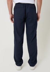 Persona vistiendo pantalones casuales azul marino con cintura elástica y bolsillos traseros, combinados con zapatillas blancas, de pie contra un fondo liso.