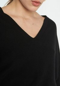 Czarny, dzianinowy sweter z dekoltem w serek i długimi rękawami, o miękkiej fakturze i luźnym kroju. Bez widocznych wzorów ani elementów metalowych.