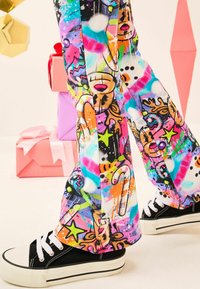 Jalat mengenakan celana bergaya grafiti berwarna-warni dengan motif liburan dan sepatu kets hitam tinggi, berdiri di dekat kotak hadiah pastel yang ditumpuk.