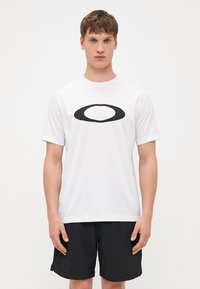 Weißes Sport-T-Shirt mit kurzen Ärmeln und einem schwarzen ovalen Logo auf der Brust. Der Stoff wirkt glatt und leicht.