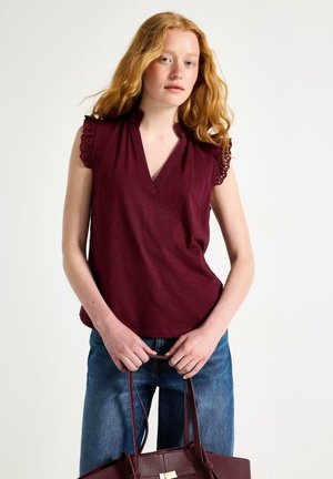 Giovane donna con capelli rossi che indossa una canotta bordeaux senza maniche con maniche di pizzo, jeans blu, tenendo le maniglie di una borsa bordeaux coordinata.