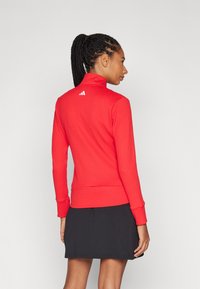 Blusa longa de mangas com textura vermelha, colarinho alto e punhos elásticos. Apresenta um logo branco nas costas. Combinada com uma saia preta.