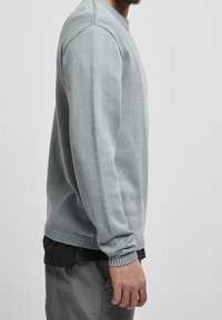 Muž v svetle šedém pleteném svetrů přes černou košili a šedé kalhoty, stojící bokem na jednoduchém světlém pozadí.