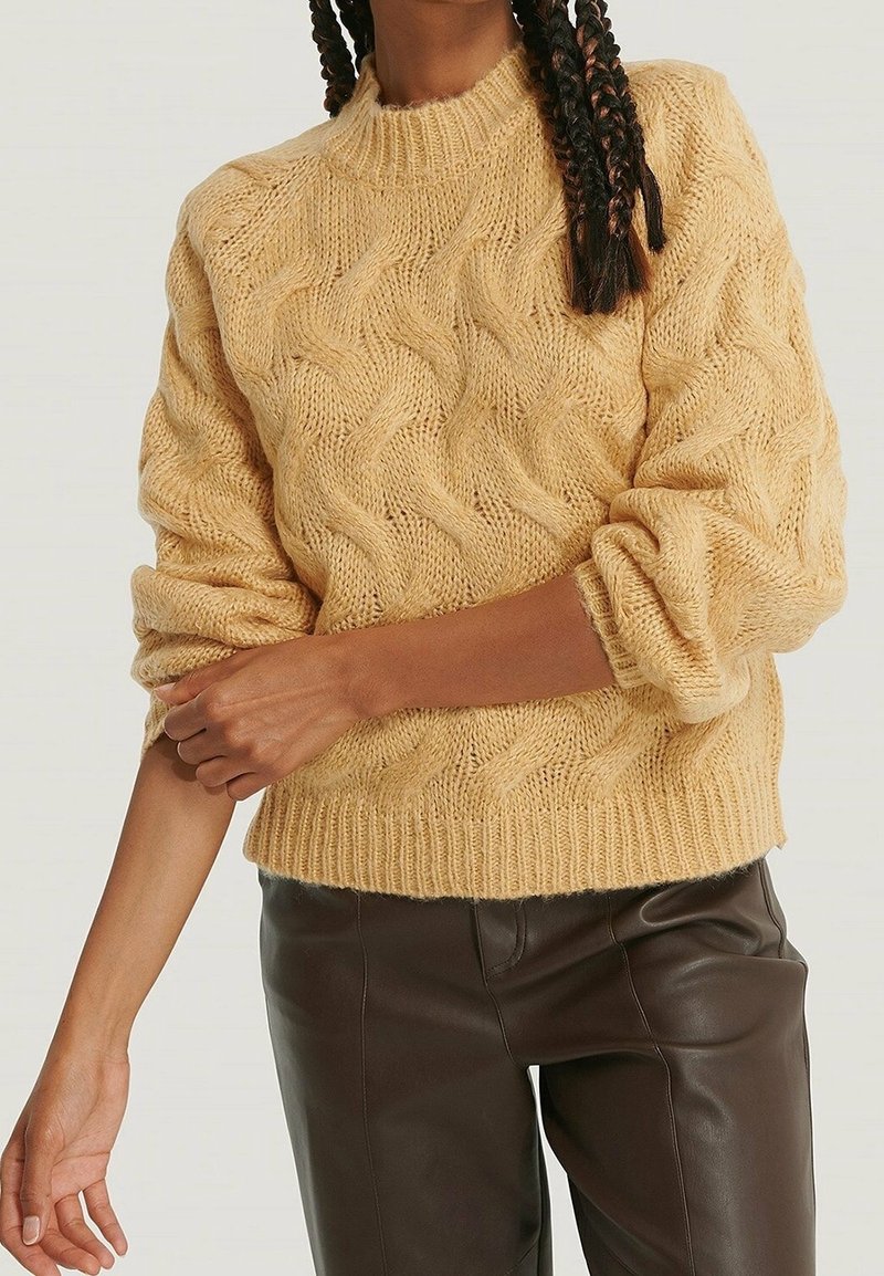 Pull en maille cablée jaune clair avec un col montant et des manches retroussées, fabriqué en matériau doux, associé à un pantalon en cuir marron.