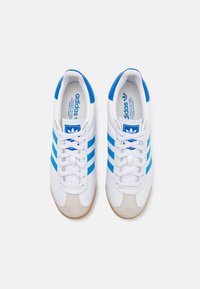 Tenisky Adidas s bielou koženou vrchnou časťou, modrými pruhmi a béžovou zamší na špičke. Nízky dizajn s bielymi šnúrkami a polstrovanou stielkou.
