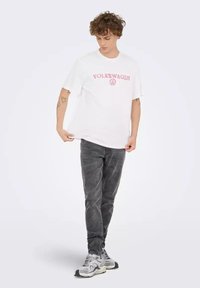Camiseta blanca con el texto y el logo de "VOLKSWAGEN" en rosa, combinada con jeans negros desgastados y zapatillas grises. Diseño casual y de corte holgado.