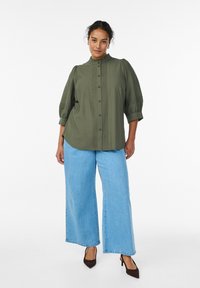 Olivgrön skjorta med knappar framtill, volangkrage, veckad front och voluminösa ärmar, kombinerad med vida ljusblå jeans och bruna klackskor.