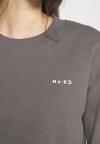 Grå sweatshirt med ribbad rundhals. Har en liten vit "NA-KD"-logotyp på vänster bröstområde. Mjuk textur, avslappnad design.