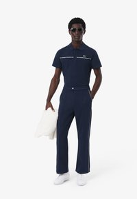 Marinblå polo skjorta med vita detaljer, matchad med marinblå byxor i skräddarsydd passform och vita sidosträck. Vita sneakers och en krämfärgad tröja.