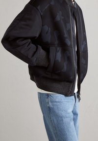 Modell som bär en svart bomberjacka med ett nätmönster och gråa ärmar, i kombination med ljusblå jeans framför en neutral bakgrund.