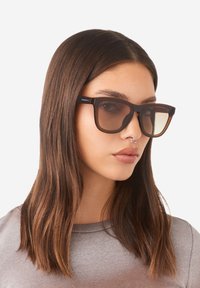 Brown oversized sunglasses with a square shape, featuring a glossy brown frame and gradient brown lenses. Minimalist logo detail on the side.