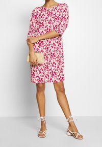 Bloemenjurk in roze en wit, korte mouwen, ontspannen pasvorm. Gecombineerd met een geweven clutch en witte sandaaltjes met dikke hakken.