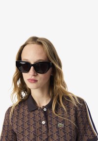 Schwarze Cat-Eye-Sonnenbrille mit glänzendem Finish, getragen mit einem braunen Polohemd mit Muster und grünem Logo. Die Haare sind wellig und locker.