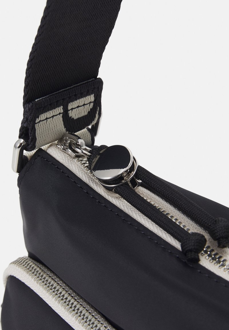 Schwarze Stofftasche mit zwei silbernen Reißverschlüssen, ausgestattet mit einem strukturierten Tragegurt und kontrastierenden, hellen Reißverschlussakzenten. Glatte und strapazierfähige Materialien.