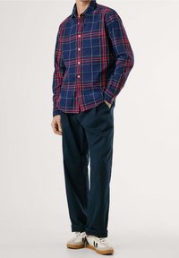 Mężczyzna w granatowej, czerwonej i białej koszuli w kratę z guzikami, w ciemnoniebieskich spodniach i beżowych trampkach z czarnymi akcentami.