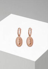 Orecchini a cerchio in oro rosa con pendenti ovali caratterizzati da dettagliata incisione e una bordatura testurizzata, esposti su una piattaforma geometrica bianca e grigia.